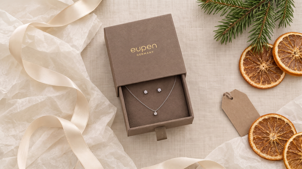 Geöffnete eupen-Schmuckschachtel mit Collier und Ohrsteckern in Weißgold mit Diamant, umgeben von Geschenkband, Tannenzweig und Orangenscheiben